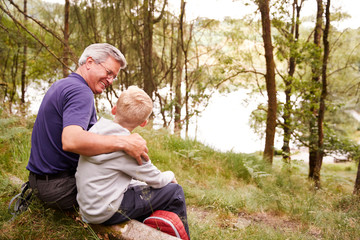 Grandfather and grandson on a hike sitting on a fallen tree in a forest, looking at each other, back view