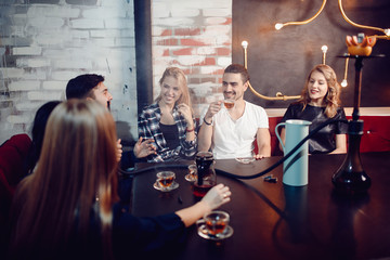 Group of friends drink tea and smoke hookah in bar with beautiful lamp on wall