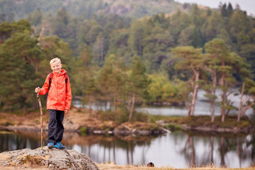 Fototapeta na wymiar A boy standing on a rock by a lake holding a stick, smiling to camera, Lake District, UK