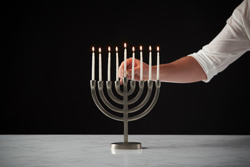 Hand Placing Lit Candle On Metal Hanukkah Menorah On Marble Surface Against Black Studio Background
