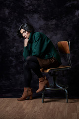 Fototapeta na wymiar Portrait of a Latin woman in Autumn-Winter outfit sitting on a vintage chair in front of a black textured backdrop.