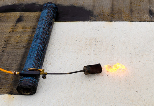 Material and equipment for waterproofing roofs and terraces. Roll of bituminous sheath, propane torch and glove resting on a bituminous membrane already laid.