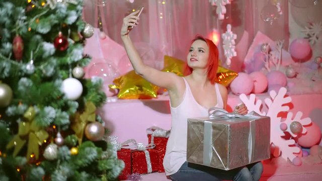 charming girl is taking selfie by camera of smartphone, sitting with big gift box in new year holiday