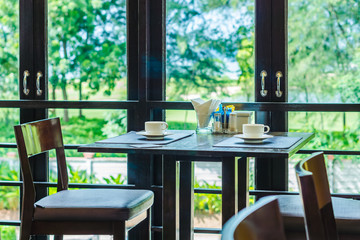 Two seats dining table sets with two coffee cups and condiments on top. Table and chairs are made of wood and They are in restaurant.