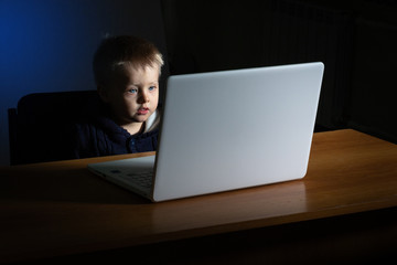 A little boy  uses a laptop in the dark.