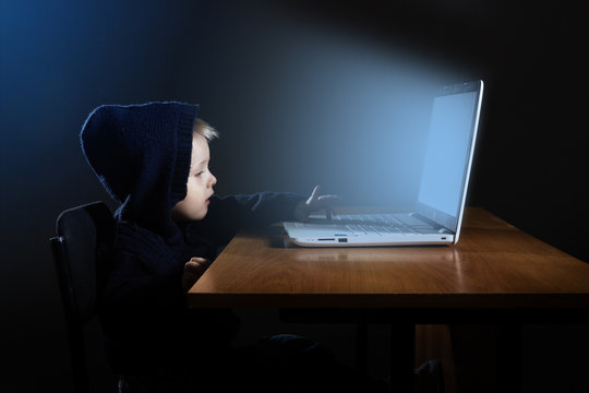A little boy in a black hood uses a laptop in the dark.