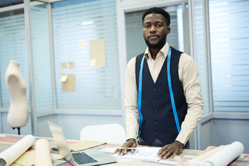 Serious confident handsome young black dressmaker with measurement tape on neck standing at table...