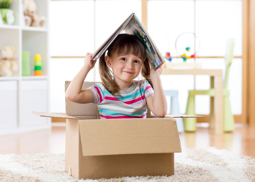 Smart kid girl sitting in box and holding a book over head as roof