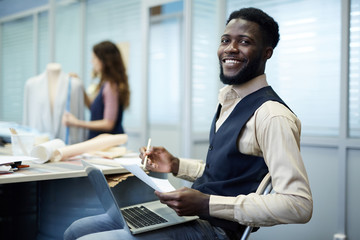 Cheerful excited young African-American fashion designer with beard sitting on chair and viewing...