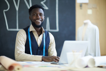 Happy young black male clothing designer with beard sitting at table and using laptop in studio, he...