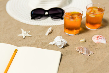 vacation, travel and summer concept - notebook with pencil, two glasses of aperitif cocktails, sun hat and sunglasses on beach sand