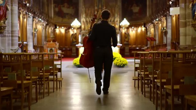 cellist man is walking in hall of catholic church with instrument, sitting on chair in front of altar