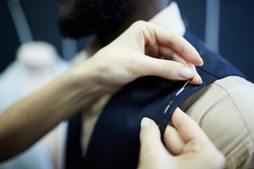 Close-up of unrecognizable tailor using sewing pins to fix fabric while correcting waistcoat during...