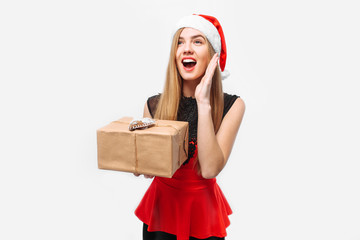 Shocked woman in a dress and Santa Claus hat, celebrating the New Year, in her hands, a Christmas gift. On a white background.