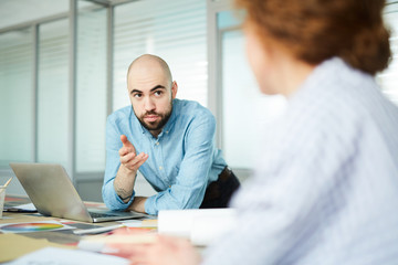 Serious bald handsome bearded designer leaning on table with open laptop and talking to colleague in modern office while they elaborating design for project