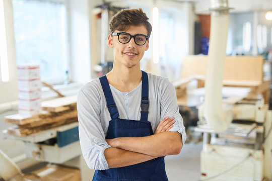 Smiling confident handsome young joiner in uniform standing in modern workshop and crossing arms on chest while looking at camera
