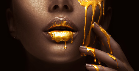 Golden paint smudges drips from the face lips and hand, golden liquid drops on beautiful model girl's mouth, creative abstract makeup. Beauty woman face