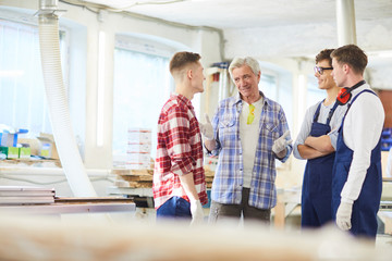 Obraz premium Smiling skilled handsome mature carpenter in checkered shirt standing in circle of young interns and explaining rules to them in modern workshop