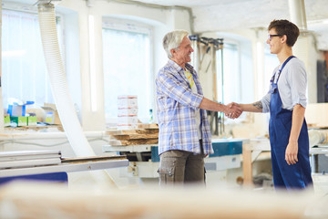 Cheerful excited mature carpenter with gray hair making handshake with young employee in overall...