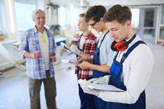Confident mature carpenter foreman explaining tasks to young workers in overalls who making notes in clipboards at practical class