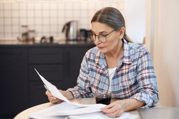 Serious aged woman in glasses checking all bills, calculating expenses. Mature housewife sitting at...