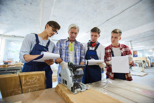 Serious concentrated woodworking specialist with gray hair standing at desk with circular saw and showing young interns with clipboards how to use it