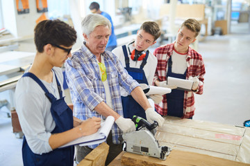 Serious experienced handsome mature carpenter in casual shirt standing at workbench and sawing...