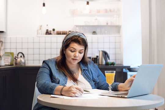 Picture of serious concentrated overweight female working through finances, sitting in kitchen with papers and open laptop on round table. People, modern technology, lifestyle and occupation