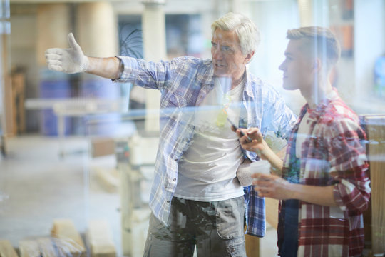 Displeased frowning mature carpenter foreman in work gloves gesturing hand and scolding young subordinate who using smartphone and drinking coffee at break