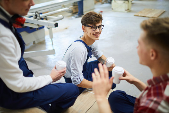 Positive handsome young workers in uniforms sitting on boxes and drinking coffee from disposable cups while having break together in workshop