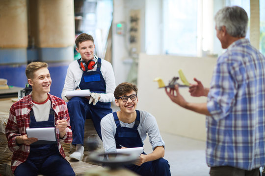 Positive excited carpentry students in overalls sitting on stack of planks and listening to carpenter during interesting class, they laughing while making notes in clipboards