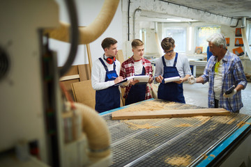 Confident skilled senior carpenter using remote control of machine while demonstrating woodworking process to young students
