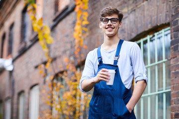 Smiling confident handsome factory worker in blue overall and glasses holding hand in pocket and looking at camera while drinking coffee outdoors