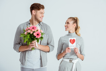 happy couple looking at each other and holding Valentines card and flower bouquet isolated on grey