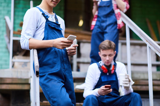 Close-up of young workers in blue overalls addicted to gadgets having break outdoors: they using smartphone and drinking coffee