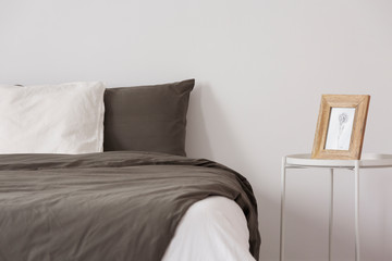 Frame on silver table next to bed with brown sheets in white simple bedroom interior. Real photo