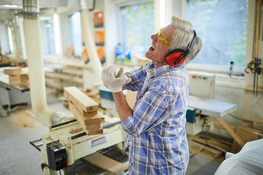 Sad emotional senior carpenter in ear protectors and safety goggles screaming from pain while hurting hand in workshop