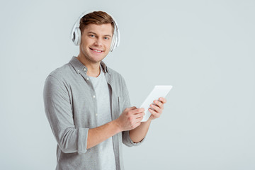 man in headphones listening music, looking at camera and using digital tablet isolated on grey