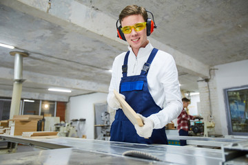 Cheerful positive handsome young carpenter with wooden board standing at desk and looking at camera...