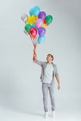 handsome man standing on tiptoe and holding bundle of colorful balloons on grey background © LIGHTFIELD STUDIOS