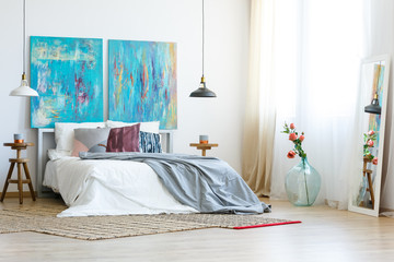 Mirror in white frame next to roses in glass vase in elegant bedroom interior with two abstract blue paintings as headboard of comfortable king size bed