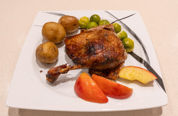 Roasted, crispy goose leg on the plate with roasted potatoes, apples and brussels sprouts.
