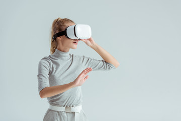 girl in grey clothing and vr headset touching air while having virtual reality experience isolated on grey