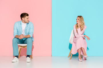 man with folded hands sitting on chair and looking at bored woman touching chin on pink and blue background
