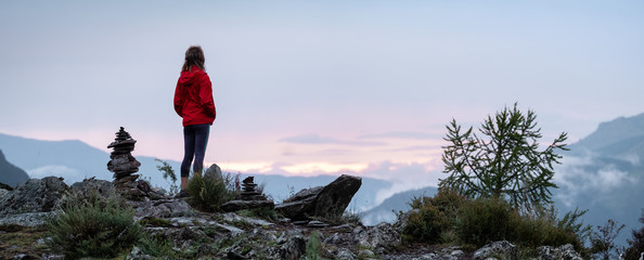 Panorama with lady hiker standing on top of the mountain among stacked stones and enjoying sunset...