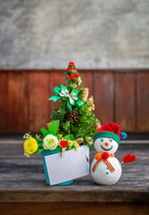 Christmas decoration item concept, Cute snowman with blank white card on flower pot over blurred Christmas tree