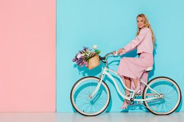 smiling woman in pink clothing riding bicycle with flower basket on pink and blue background