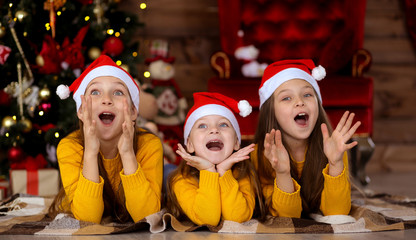 Girls in shock are smiling in red hats on the background of the Christmas tree.