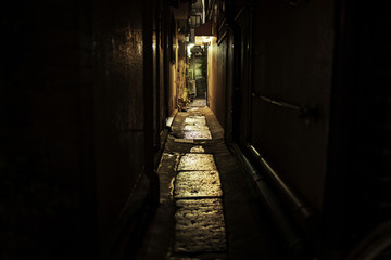 Lights shine at end of dark, narrow alley between city buildings at night