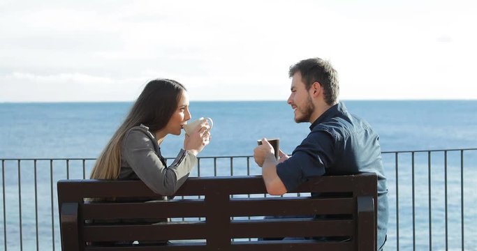 Side view portrait of a happy couple talking and drinking coffee sitting on a bench on the beach
